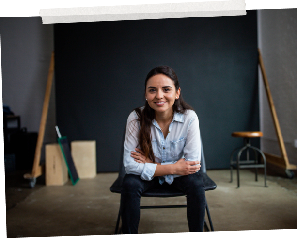 A woman sitting on a stool smiling and leaning forward with elbows resting on her legs.