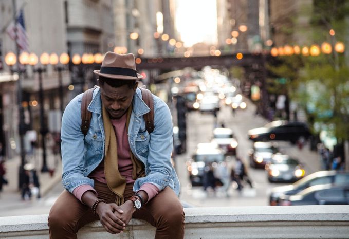 A man sitting in a hat resting his arms on his legs sitting on an overpass with a busy city street below.