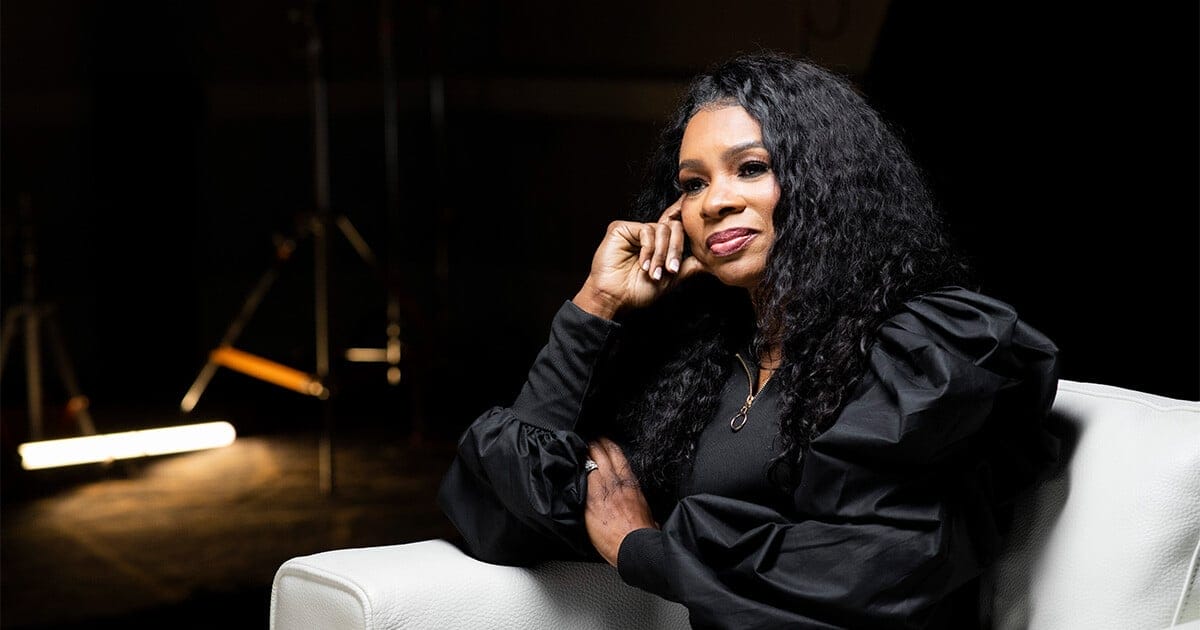 Nicole C. Mullen sitting in a white armchair looks ahead contemplating with her head resting on her right hand with her left hand across her waist.