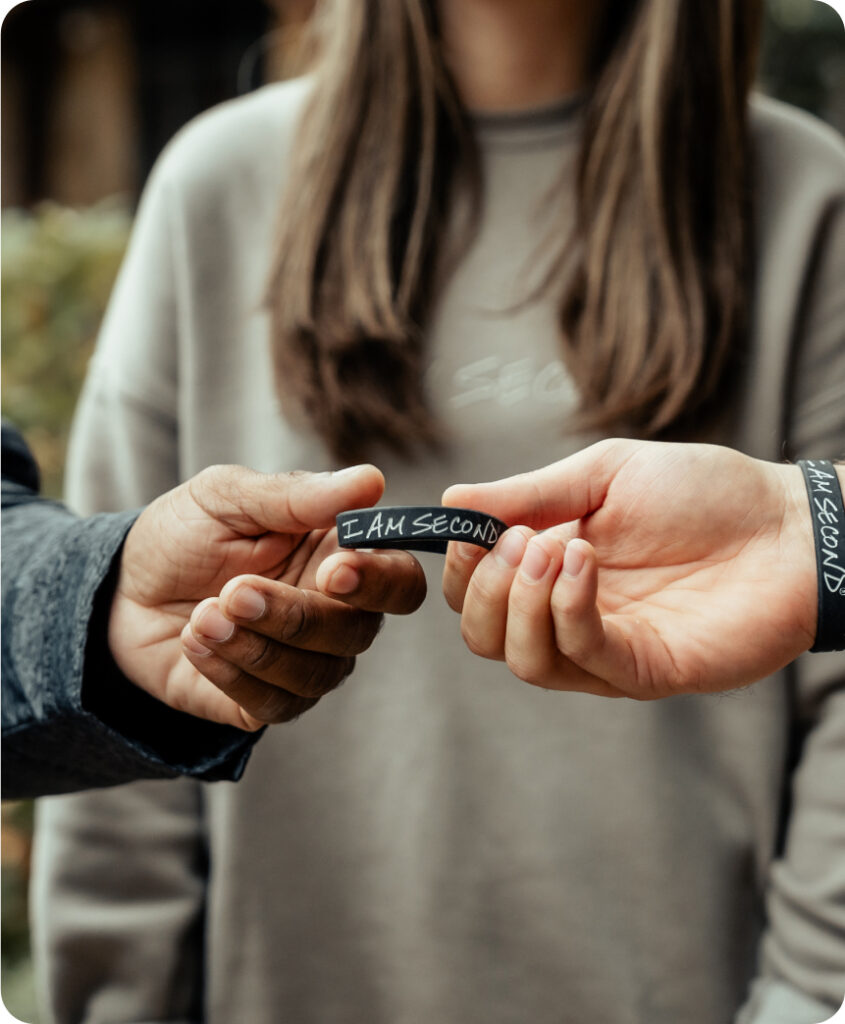 A girl stands in the background wearing an I Am Second sweatshirt while a man wearing an I Am Second wristband hands another man an I Am Second wristband.